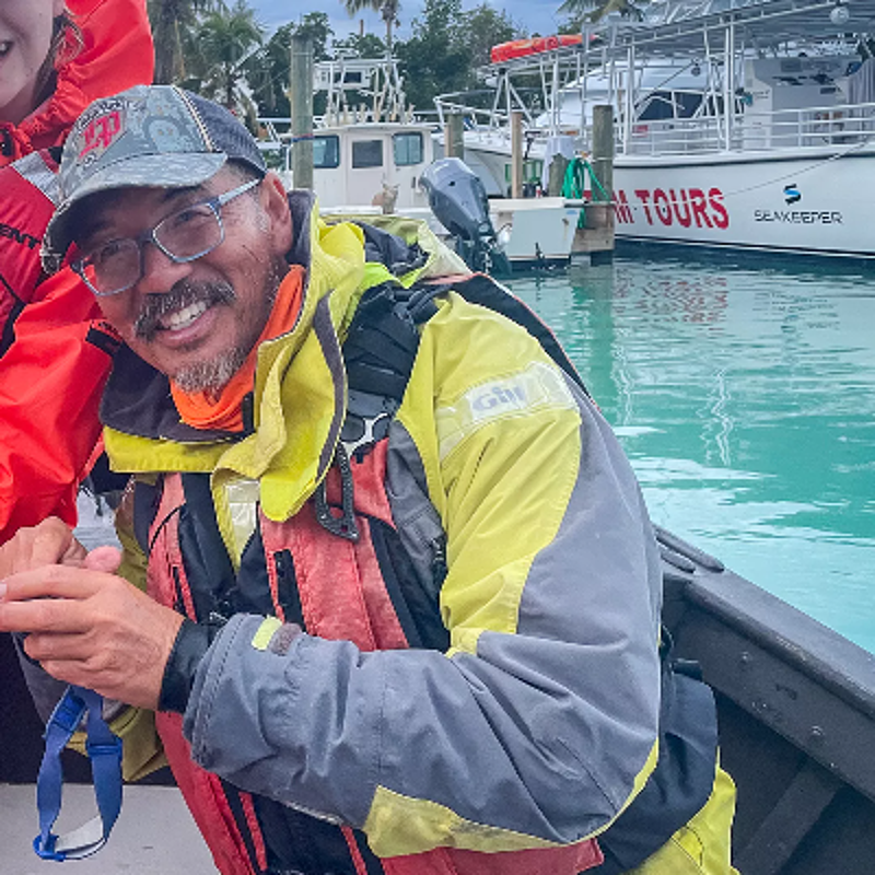 The image shows a man in a boat, wearing a hat, glasses, and a life jacket. He is smiling at the camera. In the background, there are boats and buildings. The sky is overcast.